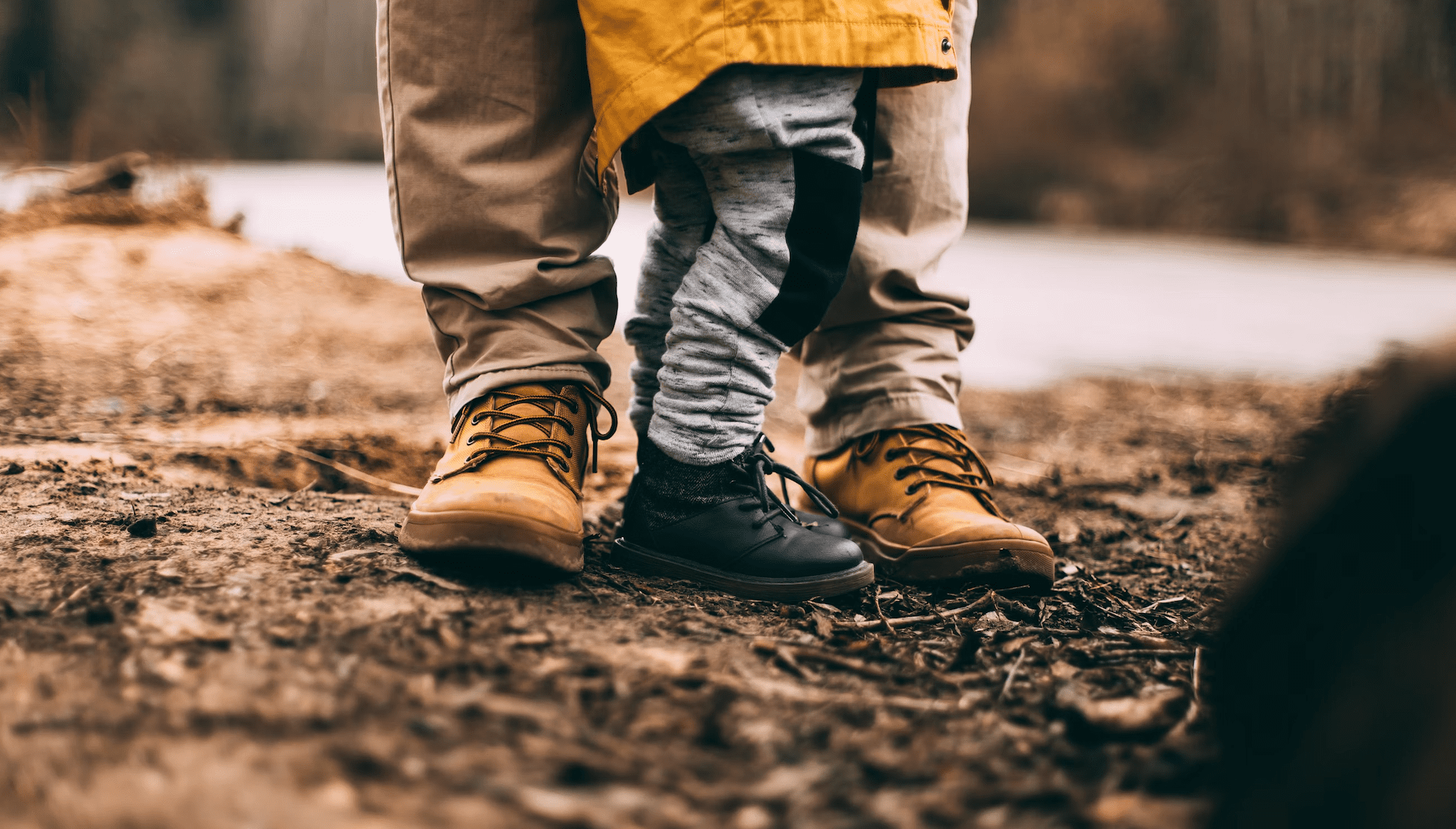 Father's boots and his child's shoes as they stand in a patch of dirt and wood chips.